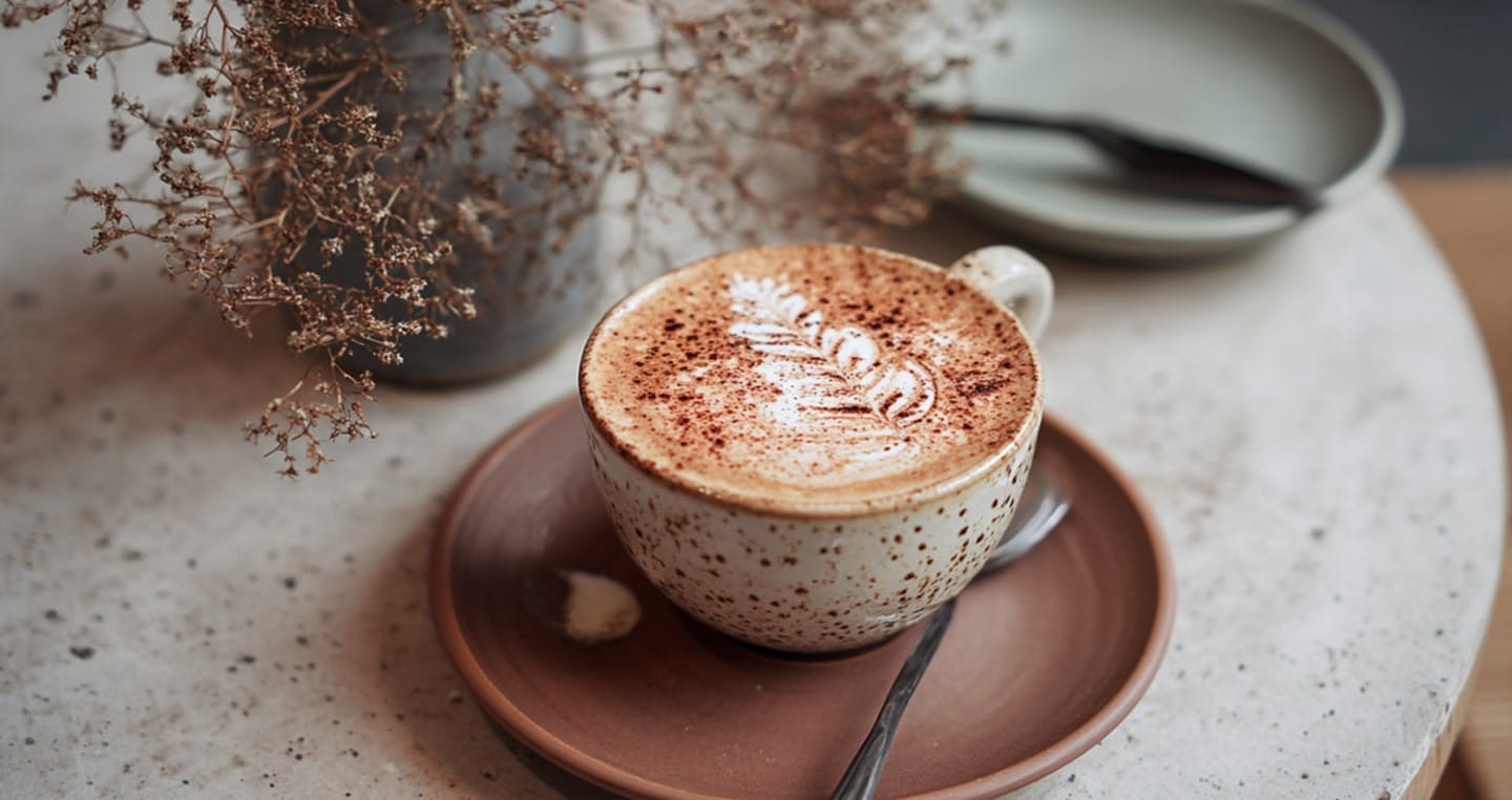 Filizanka cappuccino na talerzyku z filiżanką i suszoną rośliną w tle.