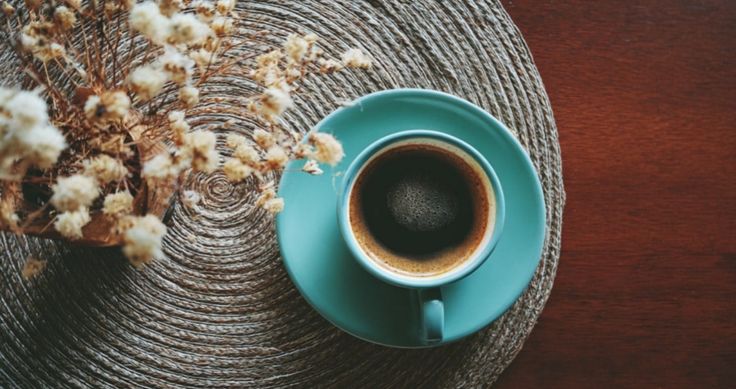 Filiżanka kawy na turkusowym spodku, obok suche kwiaty na plecionym podkładzie.