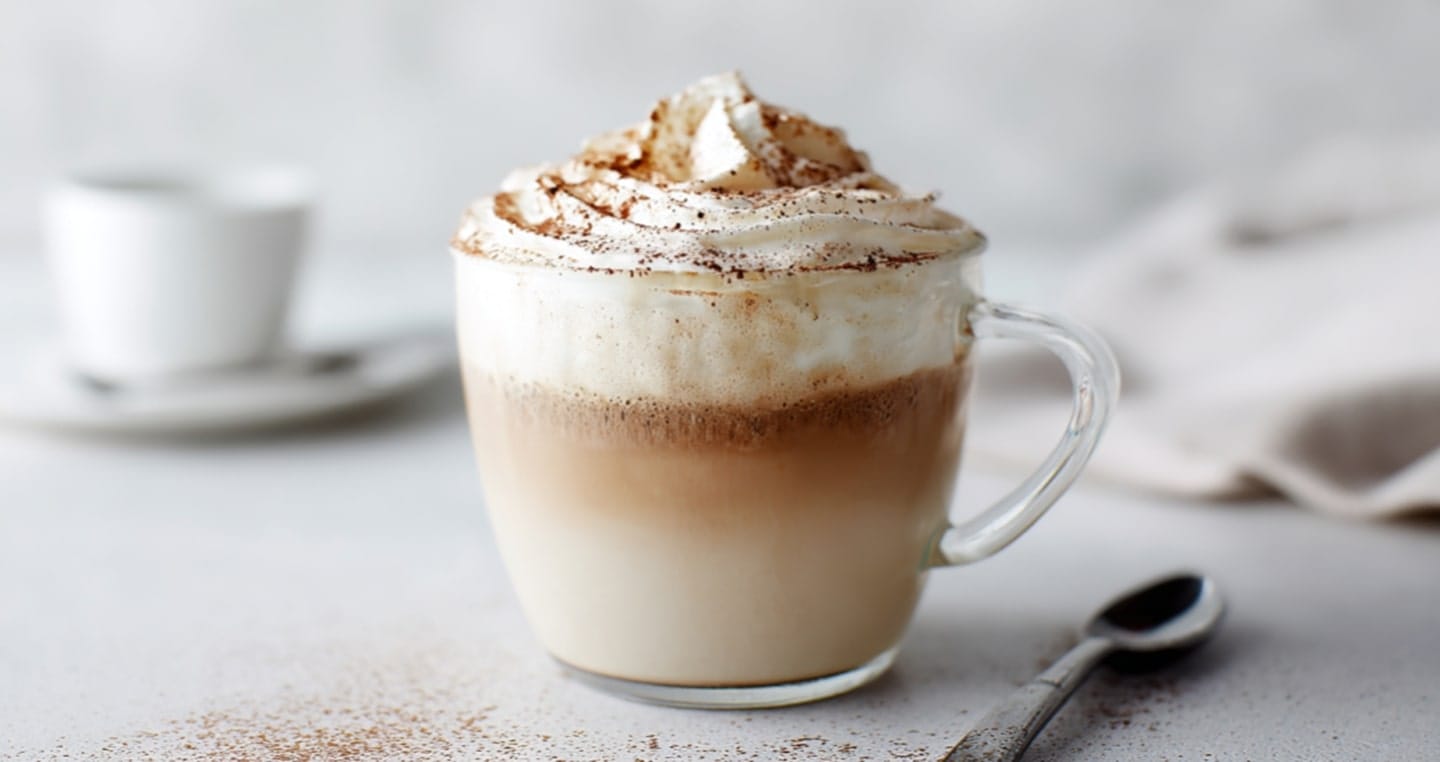 Kubek kawy latte z bitą śmietaną i cynamonem, obok łyżeczka.