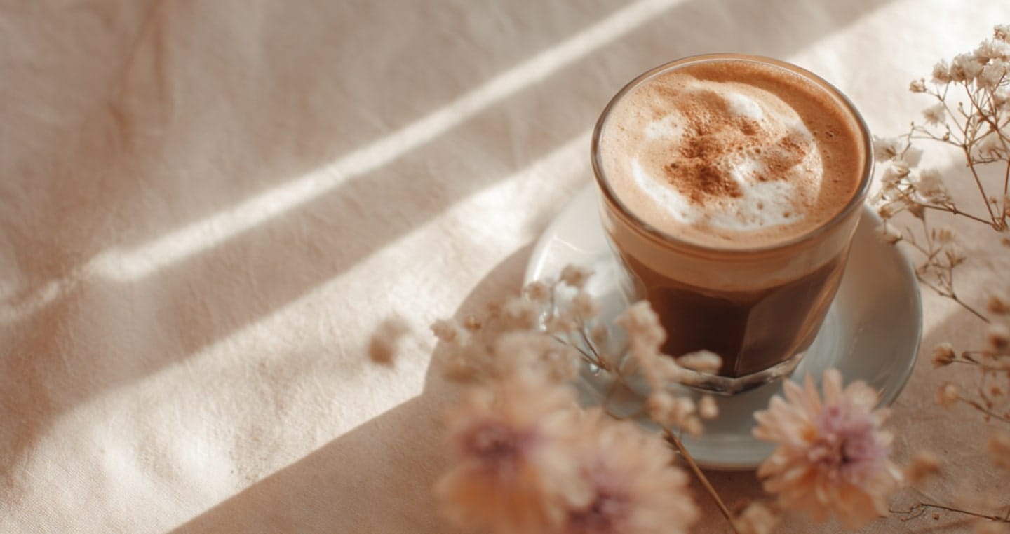 Filiżanka kawy na spodeczku, otoczona kwiatami, na tle jasnego materiału.