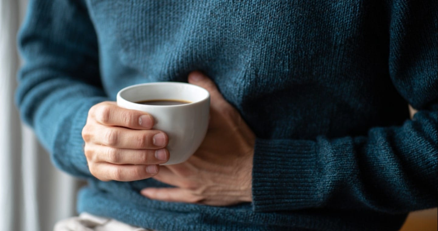 Osoba w niebieskim swetrze trzyma filiżankę kawy, ręka na brzuchu.