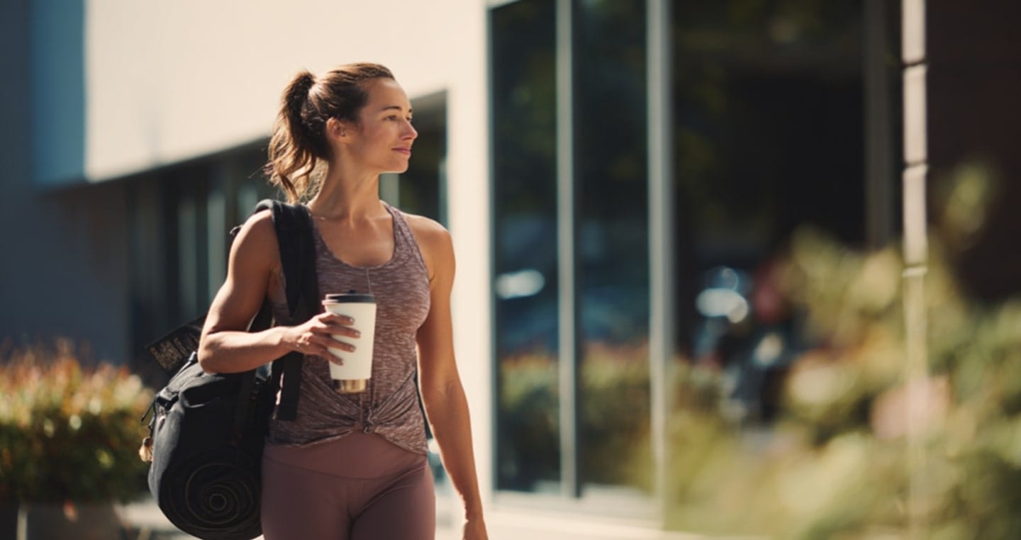 Kobieta w sportowym stroju z torbą i kawą spaceruje w słoneczny dzień.