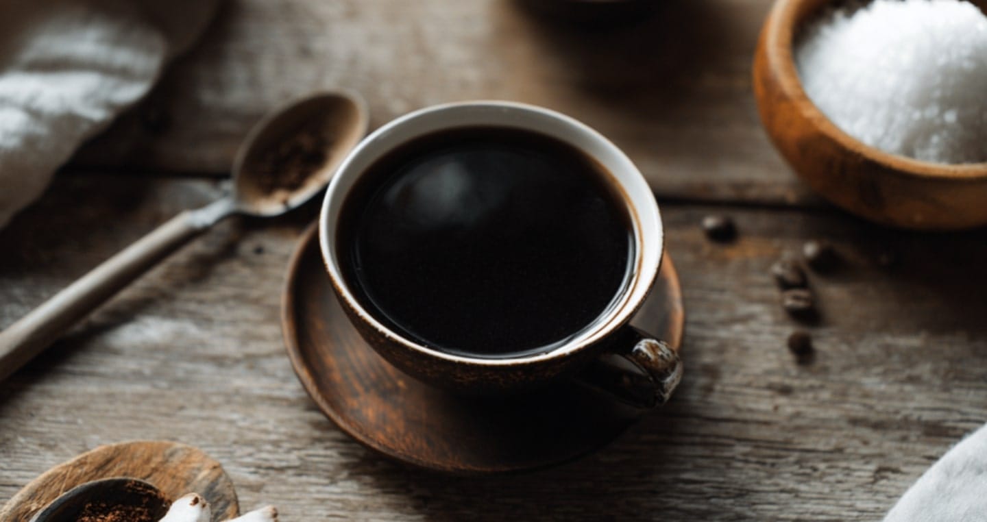 Filiżanka czarnej kawy na drewnianym stole, obok łyżka i cukierniczka.