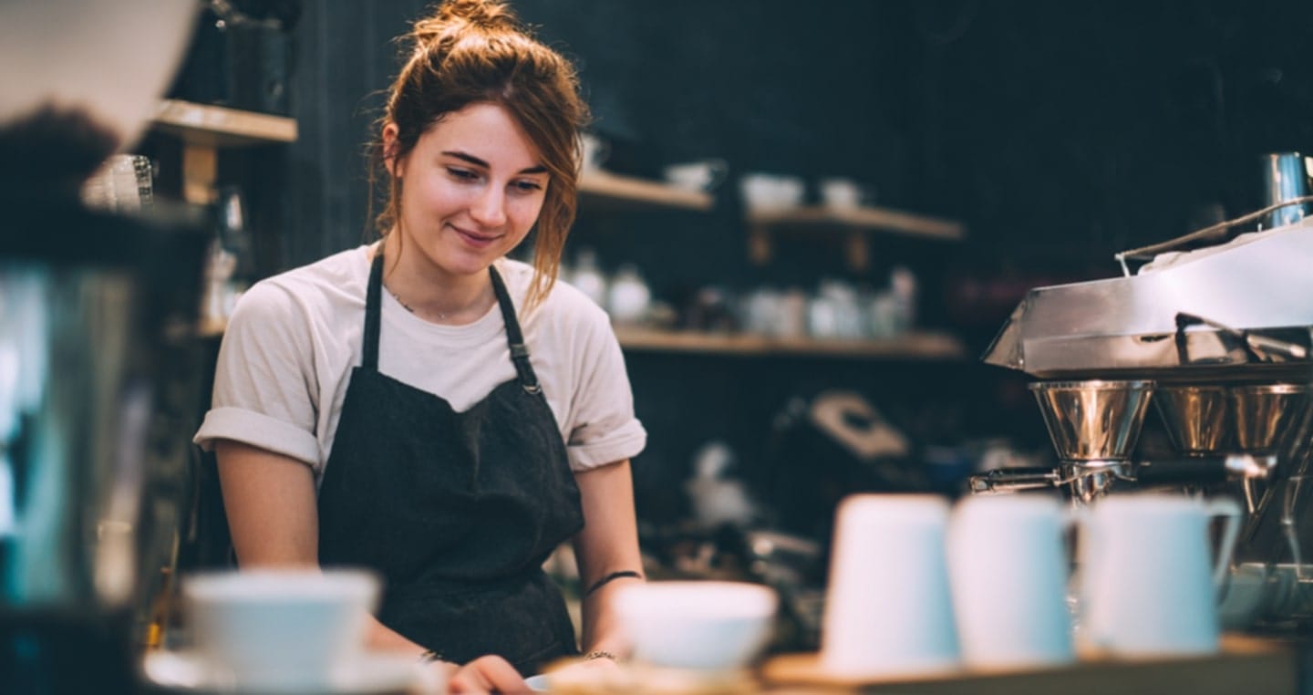 Barista w kawiarni w białej koszulce i czarnym fartuchu przy ekspresie do kawy.