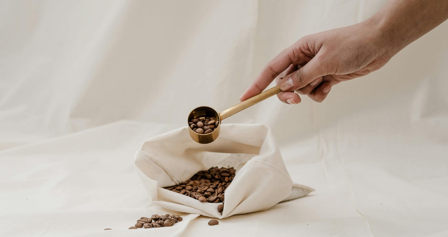 Ręka trzyma miarkę z ziarnami kawy nad lnianym workiem.
