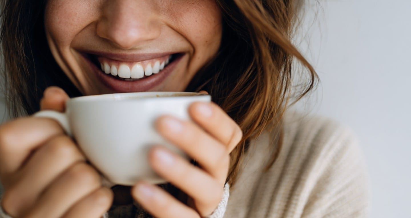 Długie, brązowe włosy. Trzymanie białej filiżanki obiema rękami. Sweter w kolorze beżowym.