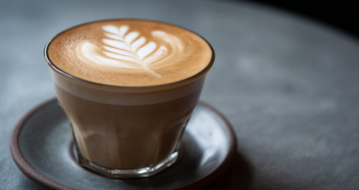 Szklanka latte z wzorem w kształcie liścia na piance, na spodku.