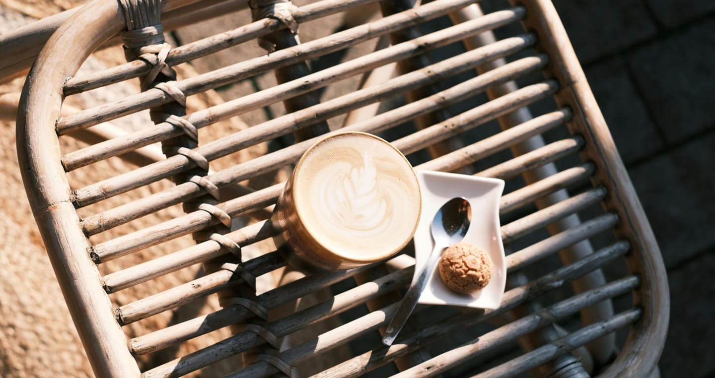 Kubek cappuccino, talerzyk z ciasteczkiem i łyżeczka na wiklinowym stole.