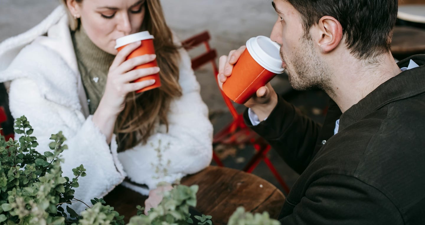Dwie osoby pijące kawę na świeżym powietrzu, w tle czerwone krzesła.