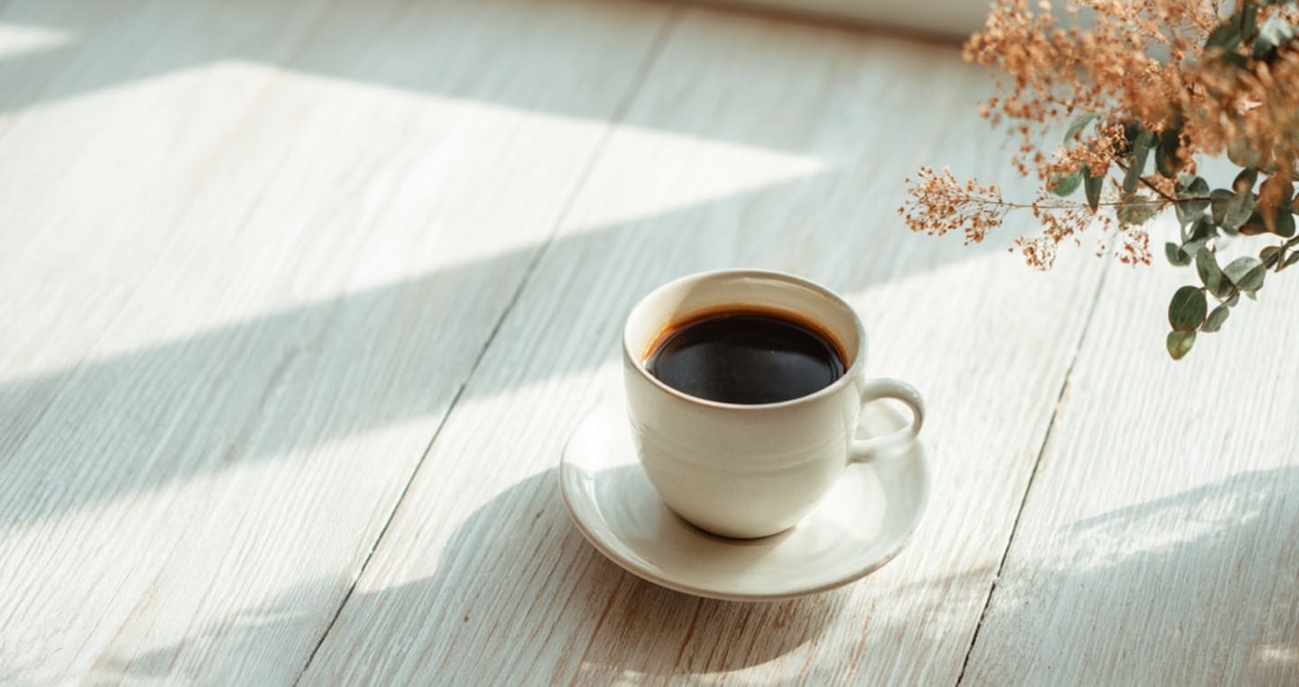 Filiżanka czarnej kawy na jasnym drewnianym stole, obok suche kwiaty.