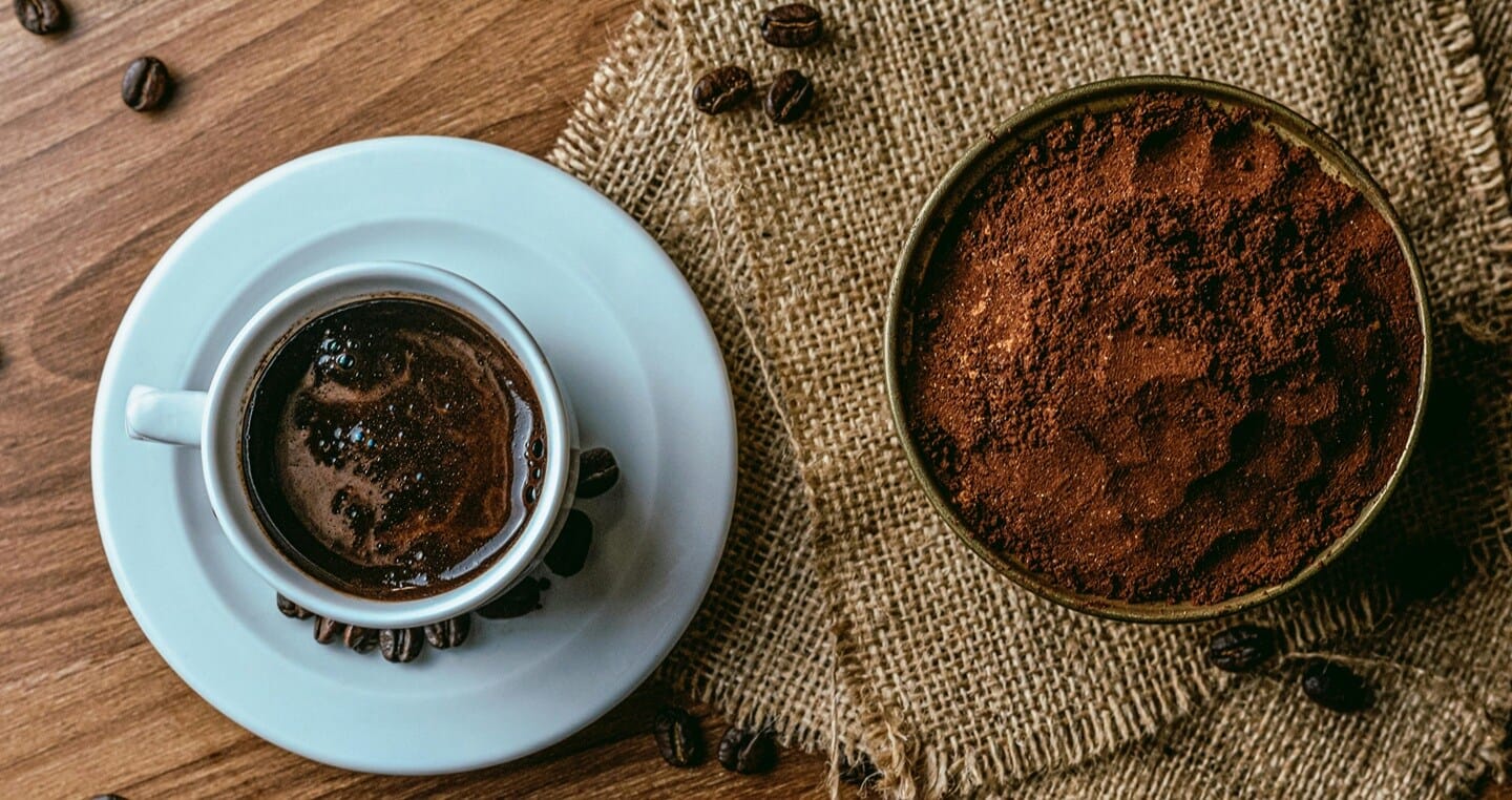 Filiżanka kawy obok miski z mieloną kawą na jutowym materiale.