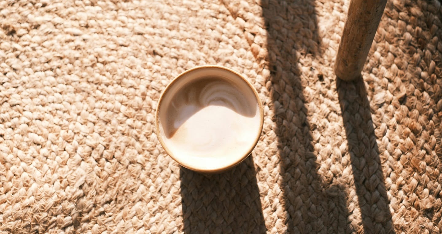 Kubek kawy na plecionym dywanie, obok nogi stołu w słonecznym świetle.