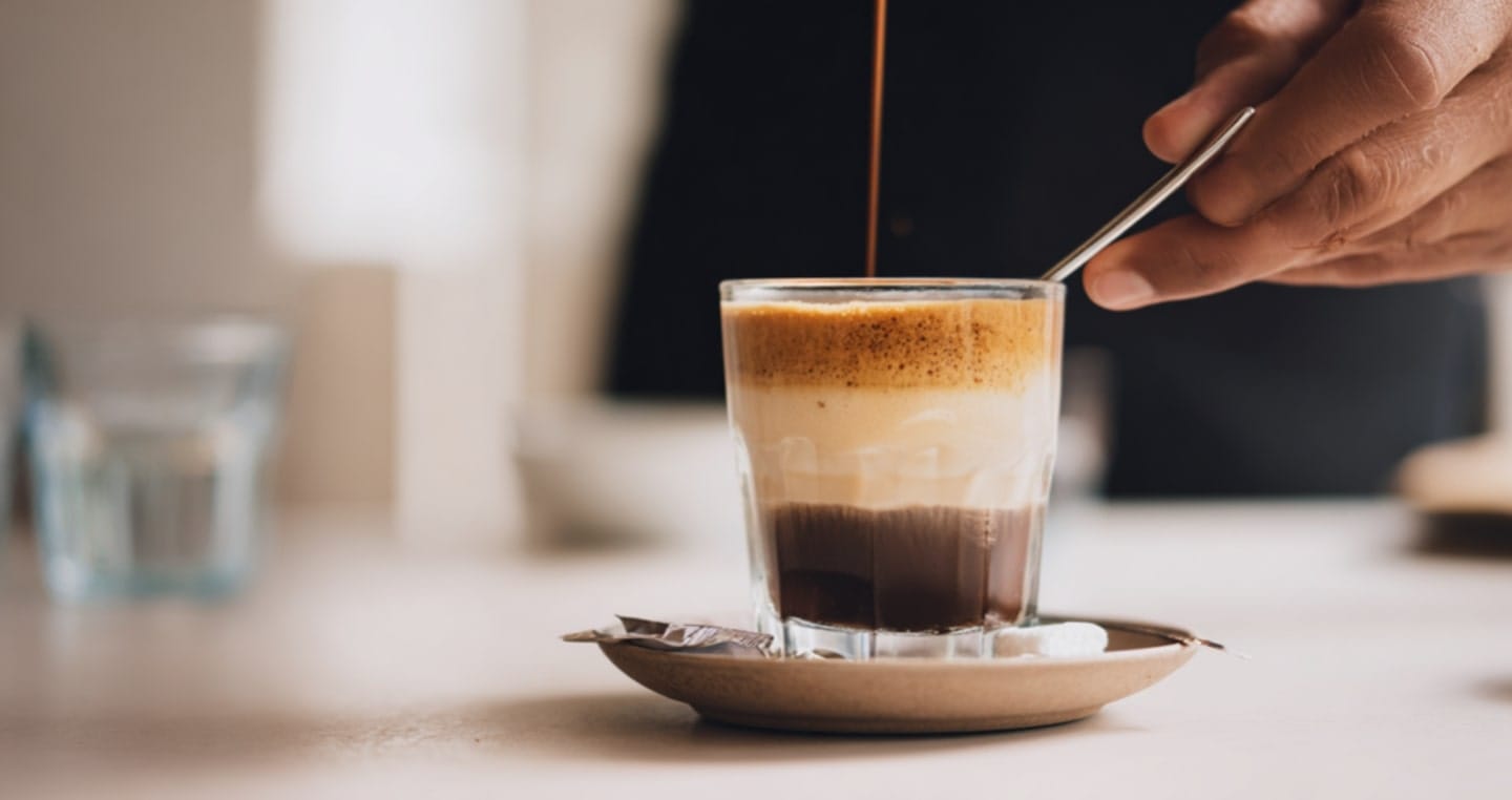 Kawa galao z mleczną pianką w szklance i ręka, która trzyma łyżeczkę.