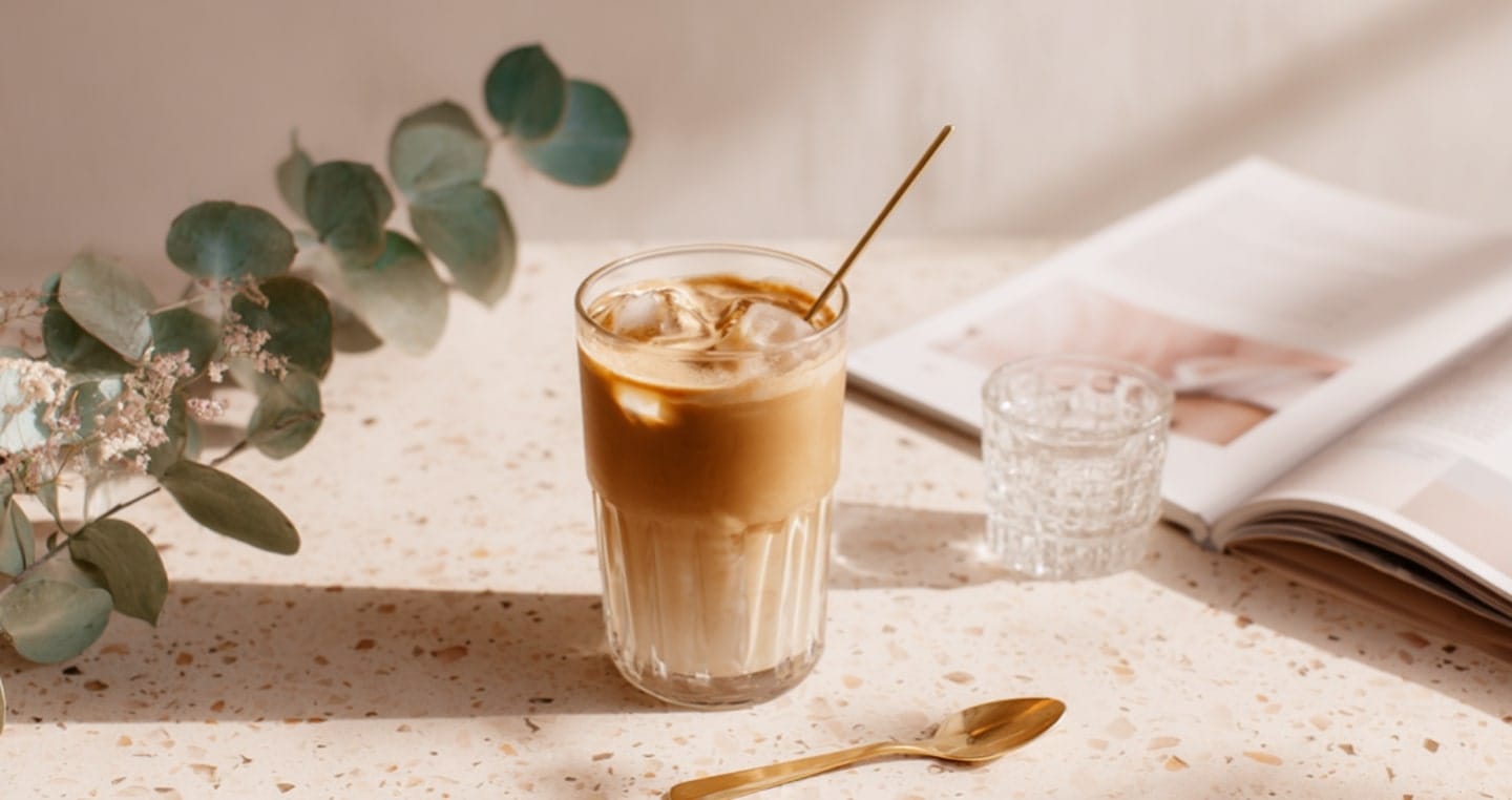 Kawa freddo, czyli szklanka kawy mrożonej z łyżką, obok roślina, książka i szklanka wody.