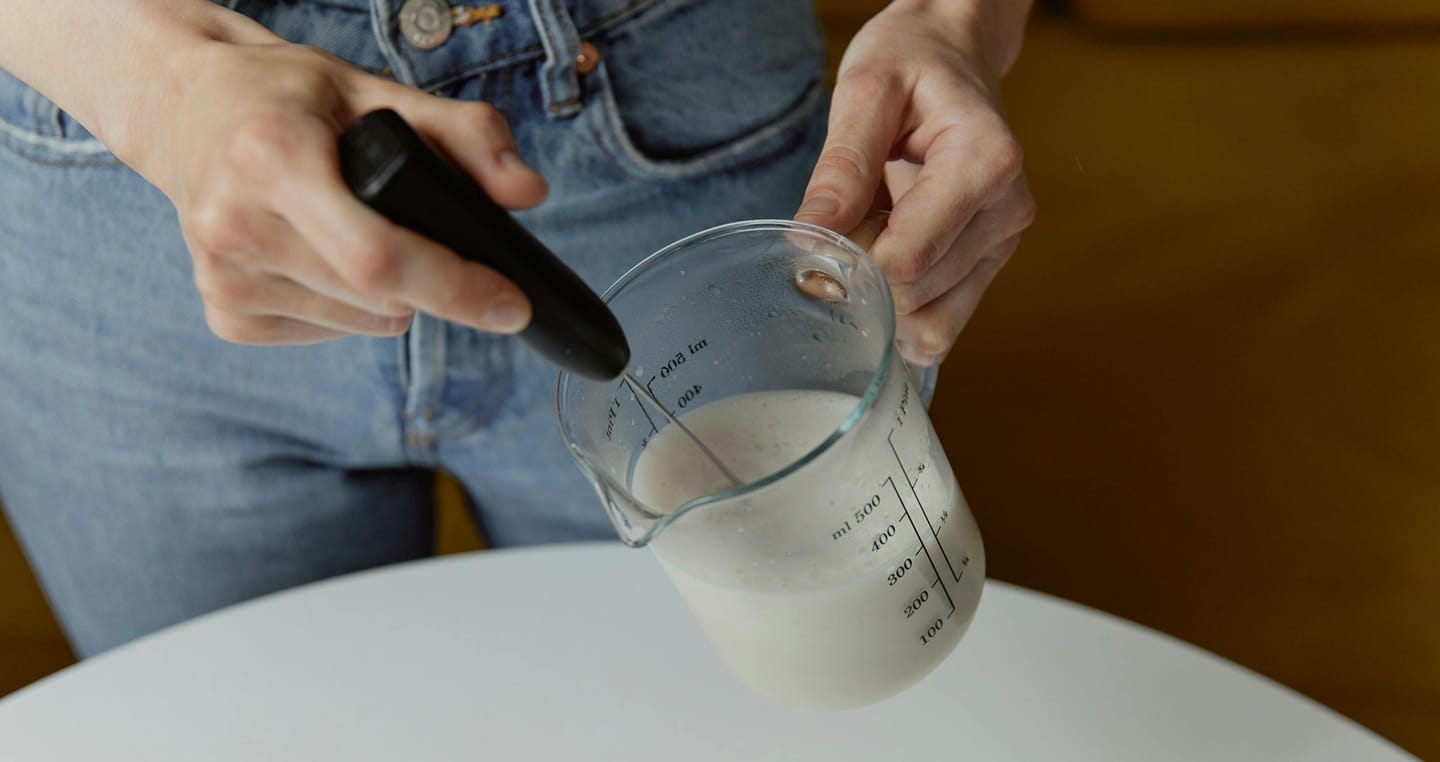 Osoba trzyma dzbanek z mlekiem i używa ręcznego spieniacza do mleka.