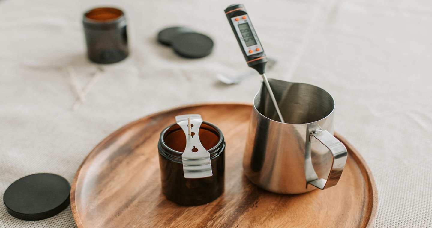 Termometr do mleka w dzbanku, słoik z kawą, pokrywki na drewnianej tacy.