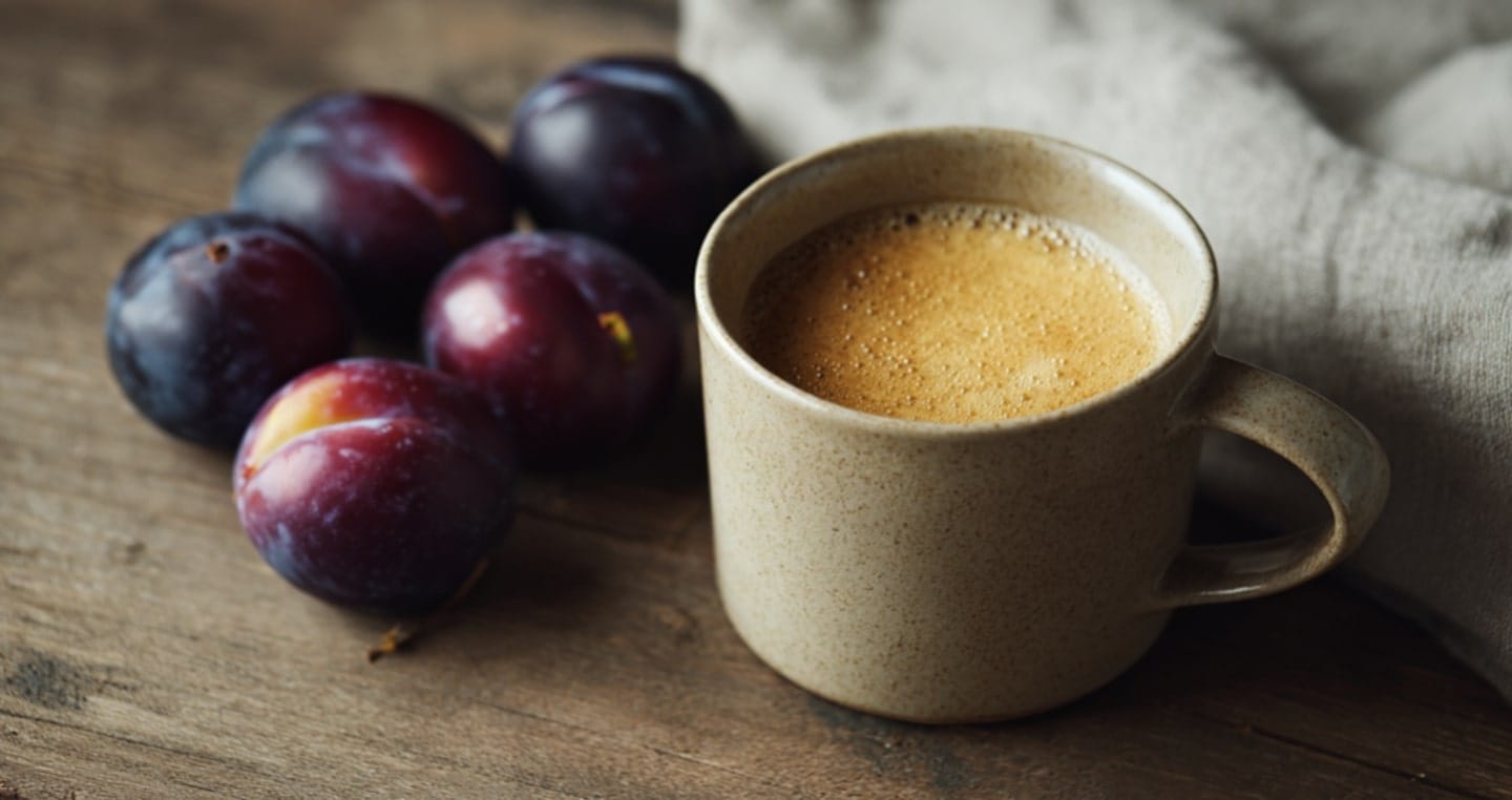 Kubek kawy śliwkowej obok trzech śliwek na drewnianym stole.