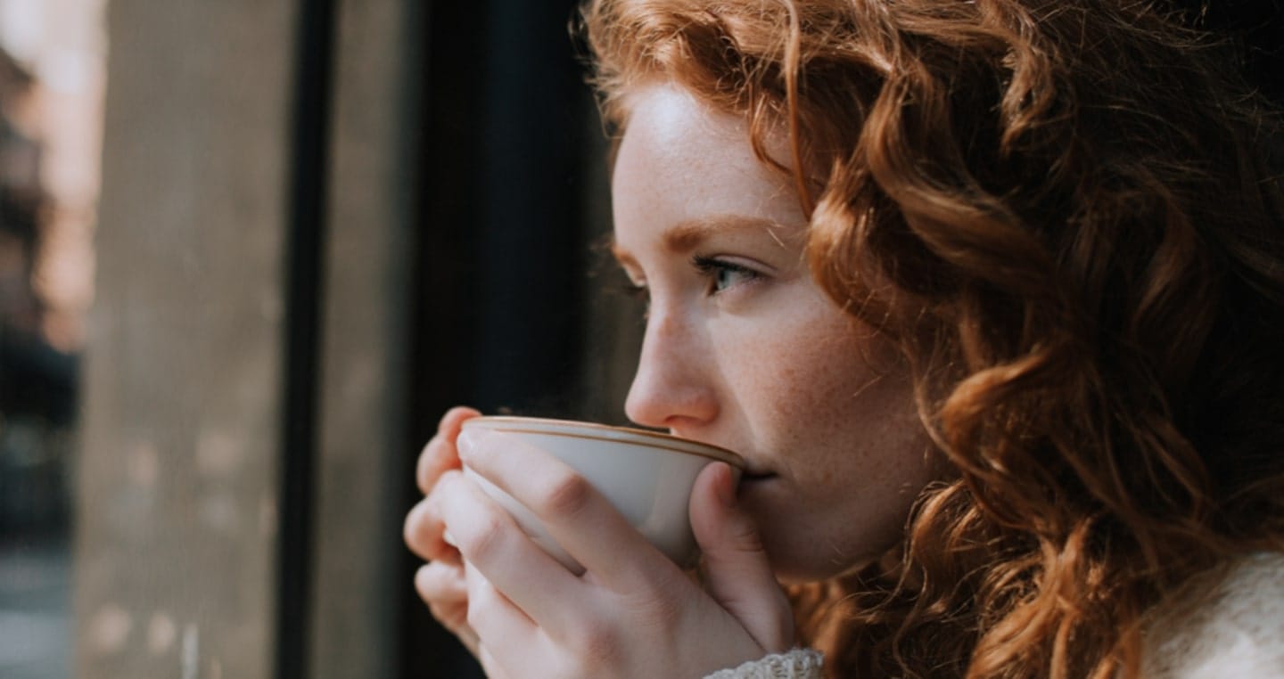 Osoba z rudymi włosami trzyma filiżankę z kawą z ashwagandhą przy oknie.
