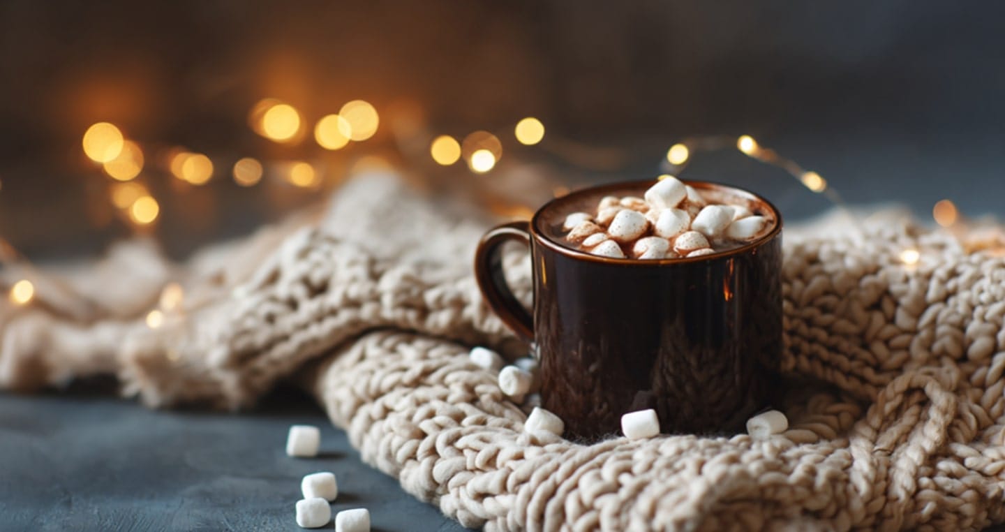 Kubek gorącej kawy z piankami na kocu, w tle światełka.