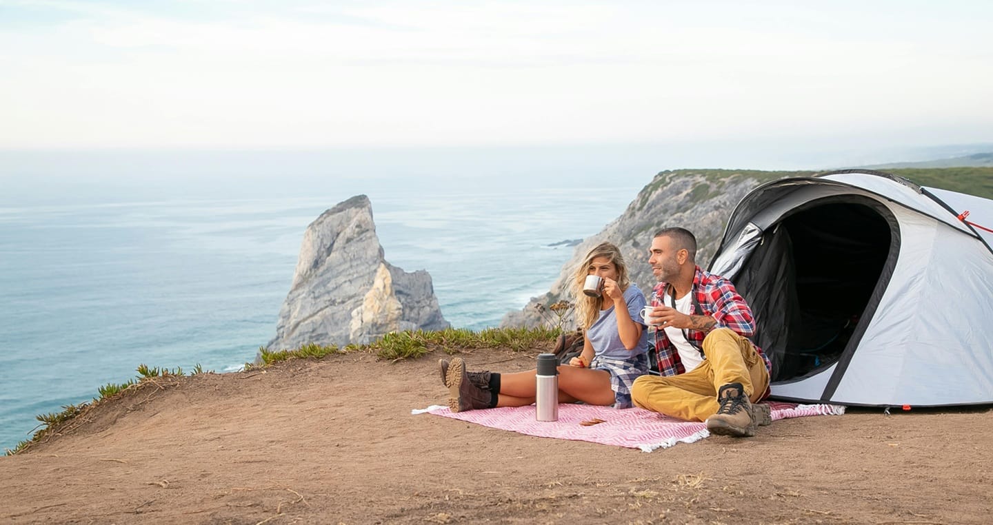 Para siedząca na kocu przed namiotem, pijąca z przenośnego ekspresu do kawy z widokiem.