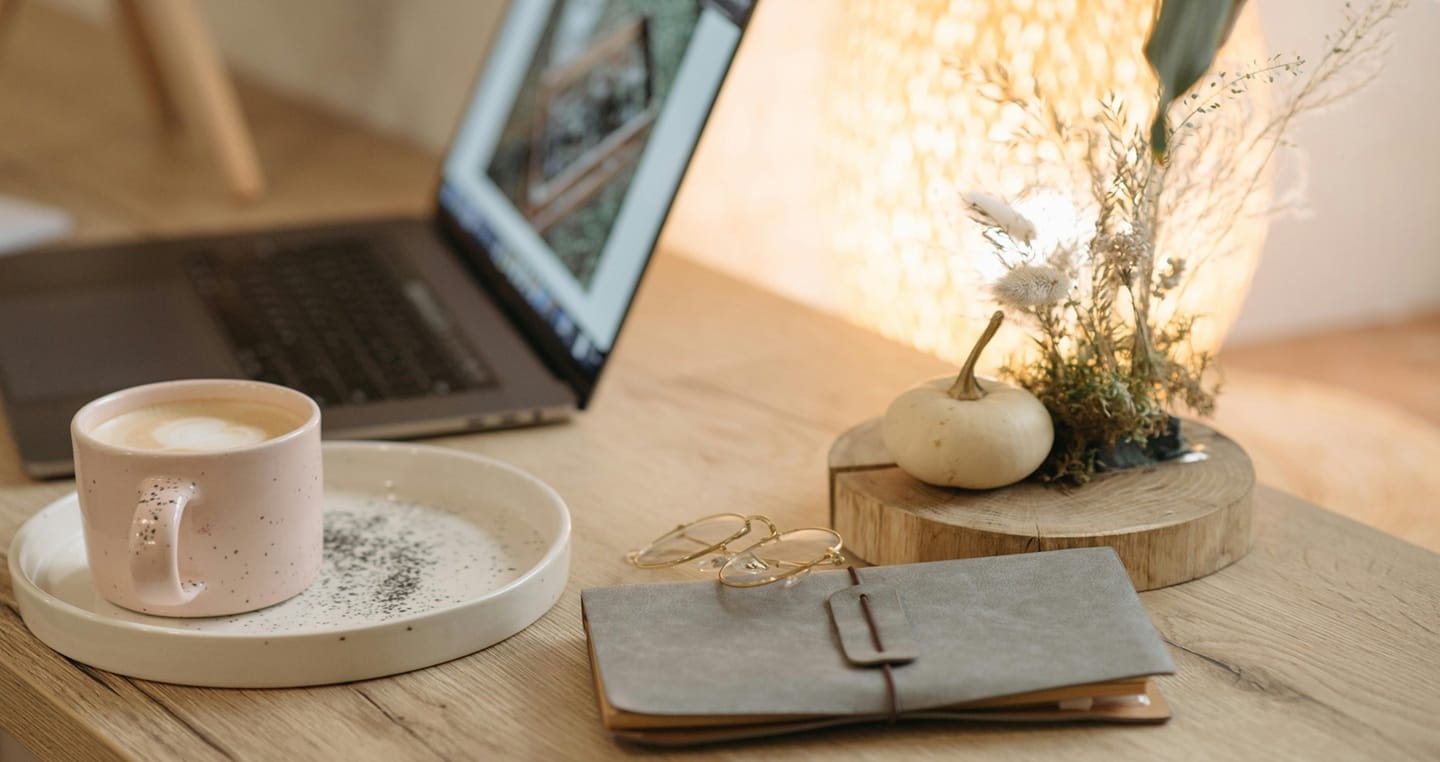 Laptop, filiżanka kawy z ekspresu tassimo, okulary, notes i dekoracja z suszonymi kwiatami na drewnianym biurku.