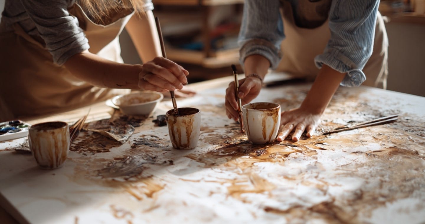 Dwóch artystów maluje na dużym stole, wykorzystując kubki z kawą.